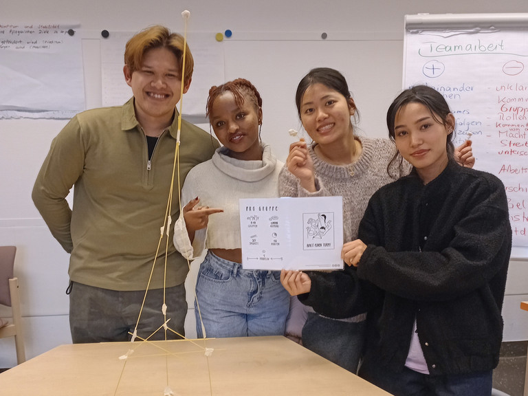 Vier Auszubildende präsentieren im Klassenraum das Ergebnis einer Teamaufgabe: einen freistehenden Turm aus Spaghetti und Marshmallows auf einem Tisch. Vier Auszubildende präsentieren im Klassenraum das Ergebnis einer Teamaufgabe: einen freistehenden Turm aus Spaghetti und Marshmallows auf einem Tisch.